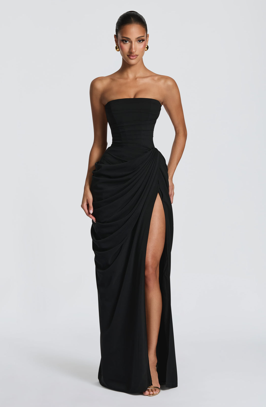 Front view of the black strapless corset dress with draped wrap skirt and high slit, one hand resting at the hip. 34