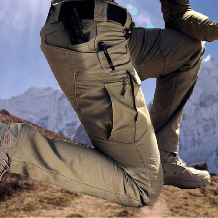 Men's beige cargo pants on a model kneeling on a mountain trail with snowy peaks behind.