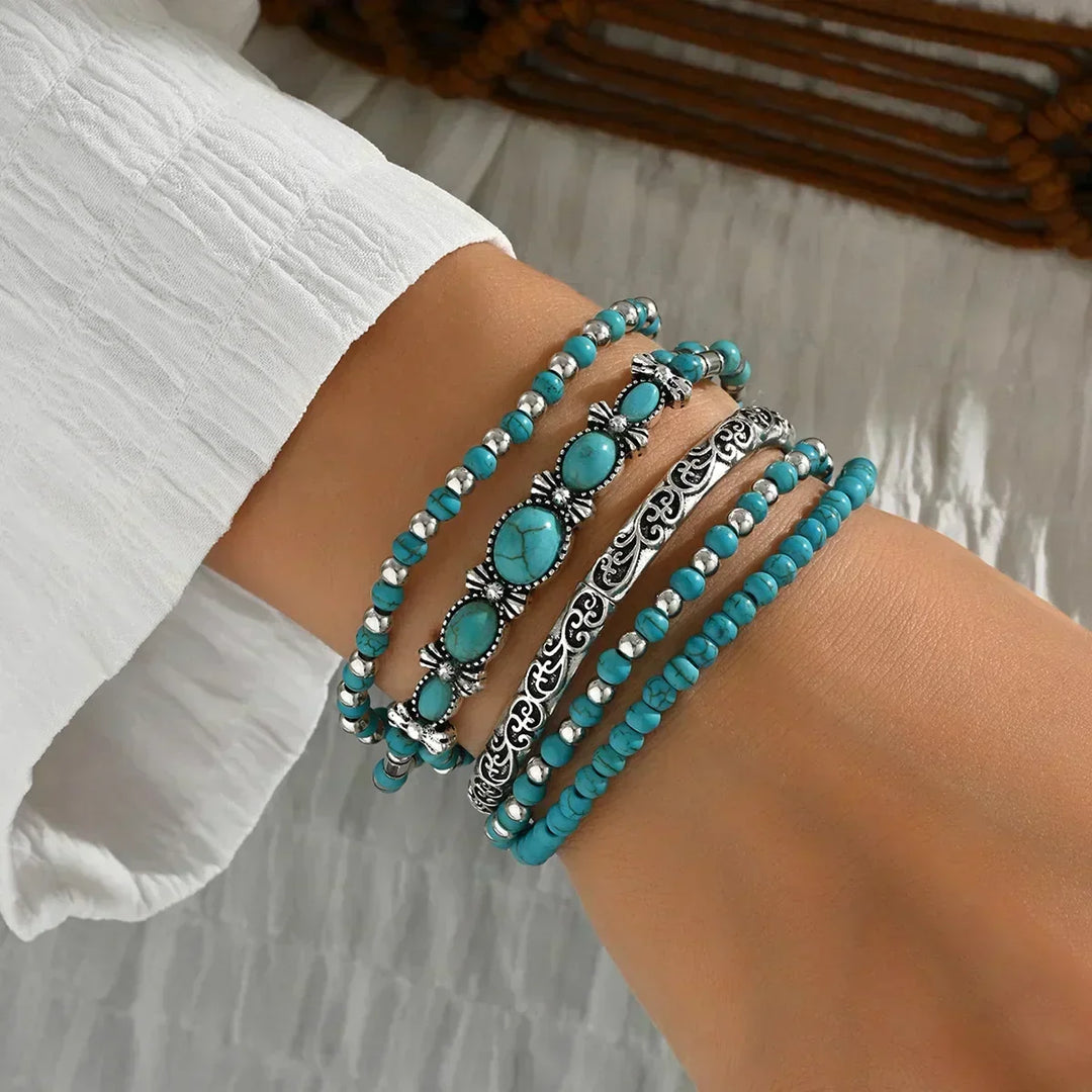 Five stacked turquoise-bead and silver-tone bracelets worn on a wrist, viewed from the side, with a white textured sleeve behind. 1