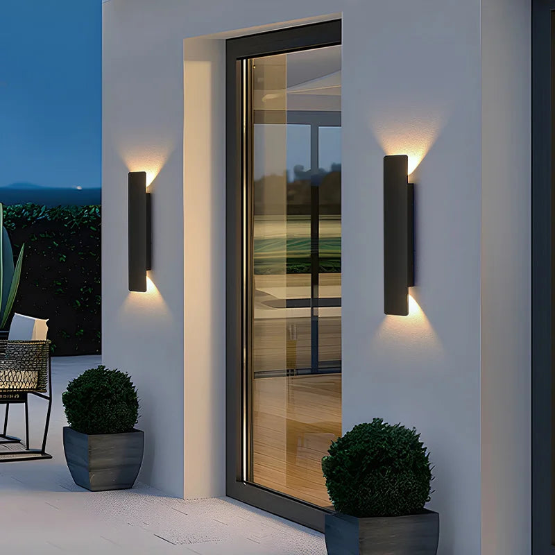Two black rectangular wall lights flanking a glass door at dusk, casting warm light up and down the white facade. 1