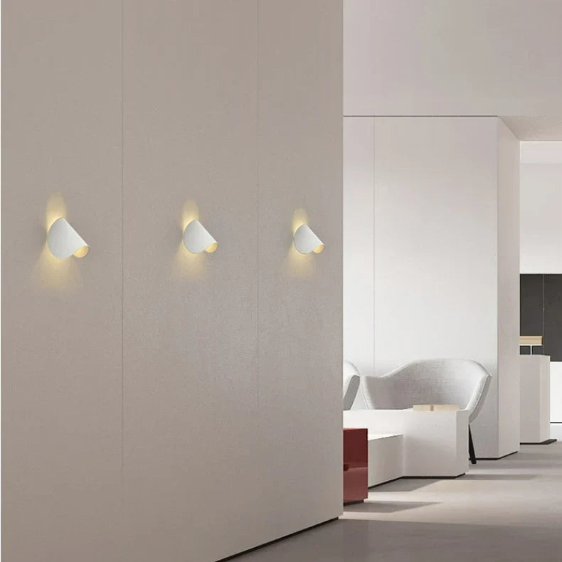Three white wall lights in a row on a long wall of a minimalist white interior, each lit warmly.