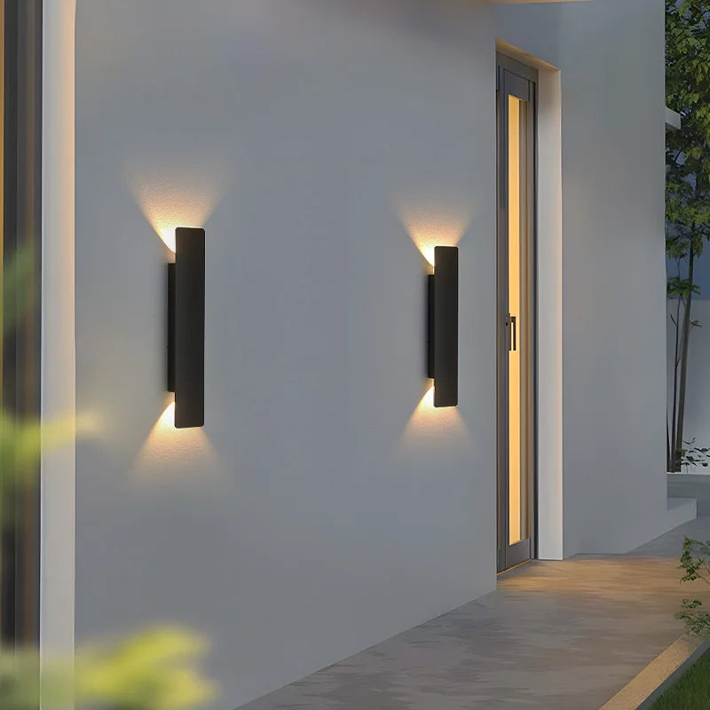 Two black wall lights on a gray exterior wall beside a tall glass door, glowing warmly at twilight. 6