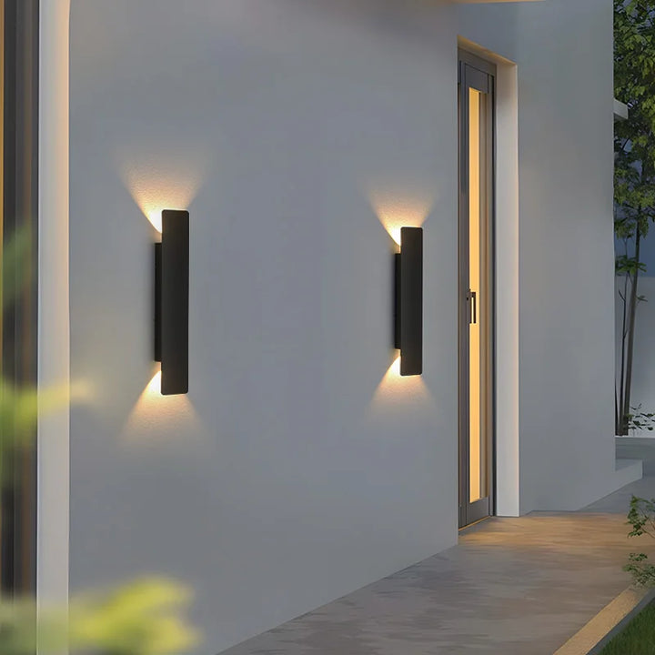 Two black wall lights on a gray exterior wall beside a tall glass door, glowing warmly at twilight. 6