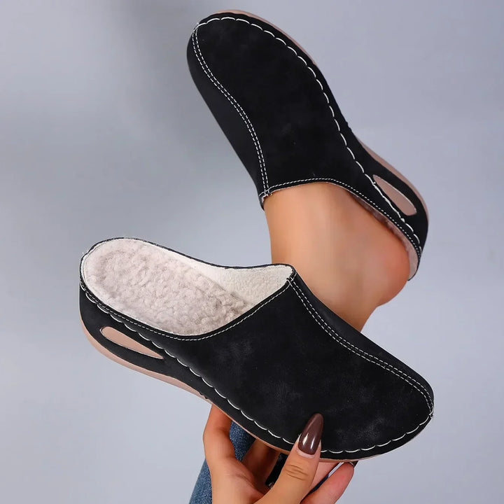 Black clog slippers held up in one hand with the cream fleece lining, white stitching and wedge sole cutout.