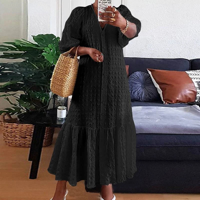 Frau in schwarzem Maxikleid mit Strohtasche vor Sofa und Pflanzen im Wohnzimmer.