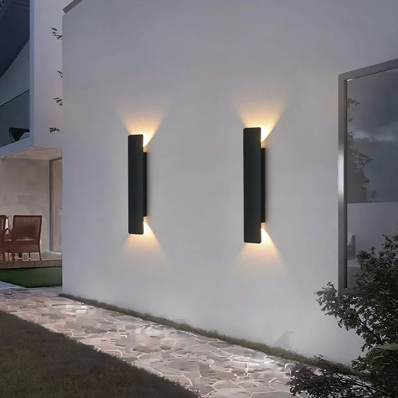 Pair of black wall lights on a long white exterior wall at night, with a stone path and lawn below. 2
