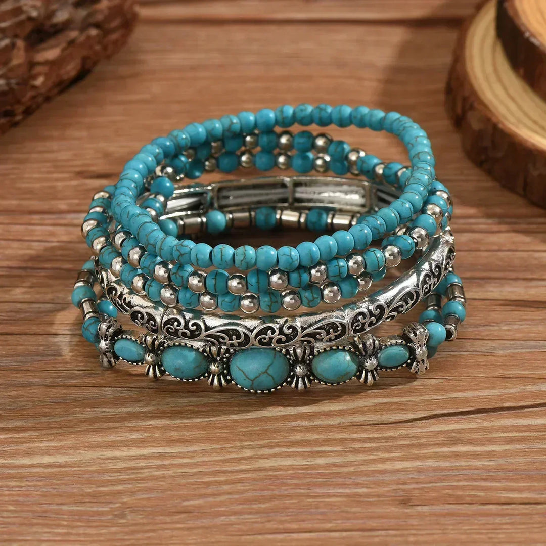 Five bracelets stacked on a wooden surface, showing the cracked turquoise beads, the carved silver-tone bangle and the oval stone bracelet.