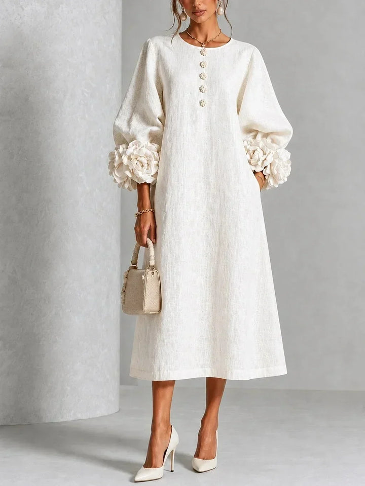 Model wears the cream white midi dress with puffed sleeves, rose-shaped cuffs and floral buttons, styled with ivory heels and a woven bag. 1