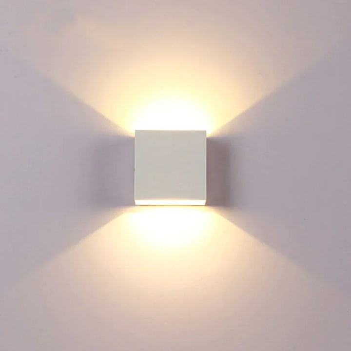 White cube wall lamp on a pale wall, shining warm light fans upward and downward. 17
