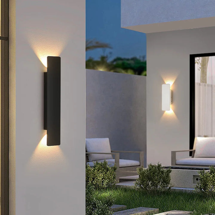 Black wall light on a patio wall at dusk, with a white version glowing in the background beside outdoor seating. 5
