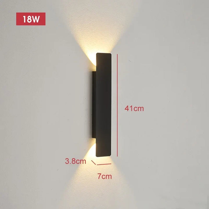 Mid-length black wall light on a pale wall casting warm light up and down, with dimension lines showing its size.