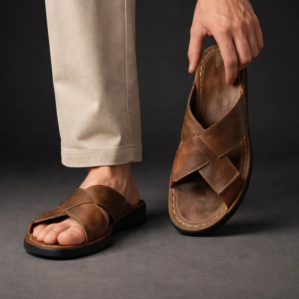 Man in beige trousers wearing one brown crossed-strap sandal while holding the other up to show its footbed. 3