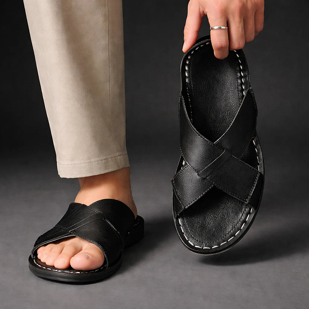 Man in beige trousers wearing one black crossed-strap sandal while holding the other up to show its stitched footbed. 7