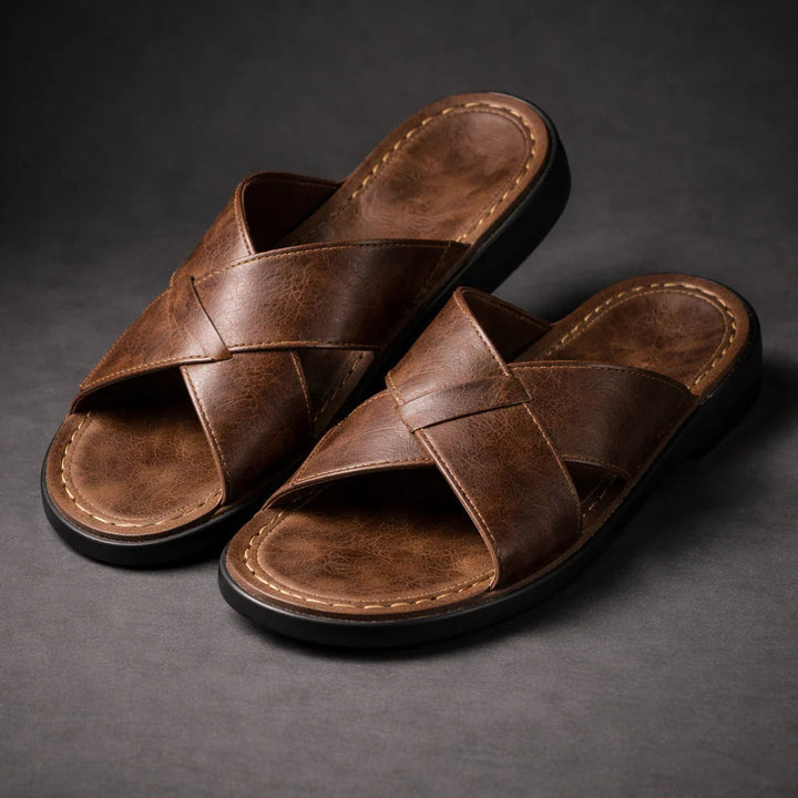 Pair of brown slip-on sandals with crossed straps and cream stitching, shown at an angle on a dark gray surface. 1