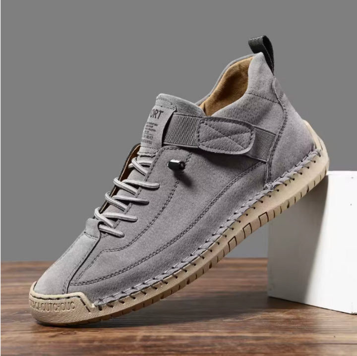 Single gray suede-look sneaker with a perforated texture, elastic strap and beige stitched sole, propped against a white block. 10