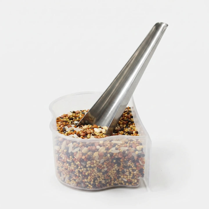 Metal scoop standing in a clear plastic cup filled with mixed seeds and grains, shown against a plain white background. 1