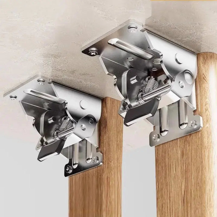Two silver metal folding leg brackets screwed under a tabletop, joining it to wooden legs, with springs and black release tabs visible. 1