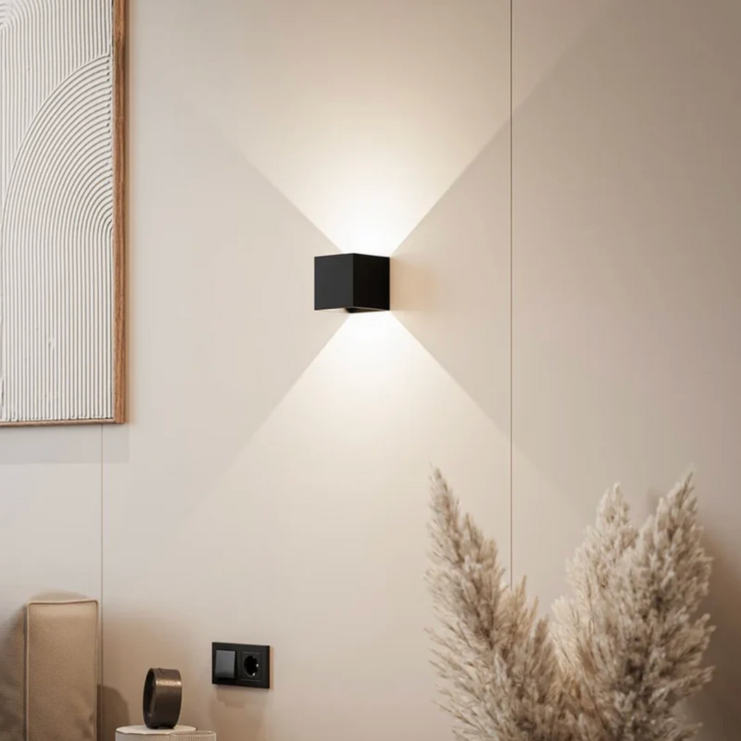 Black cube wall lamp casting warm light up and down on a beige wall beside pampas grass and a framed textured artwork. 1