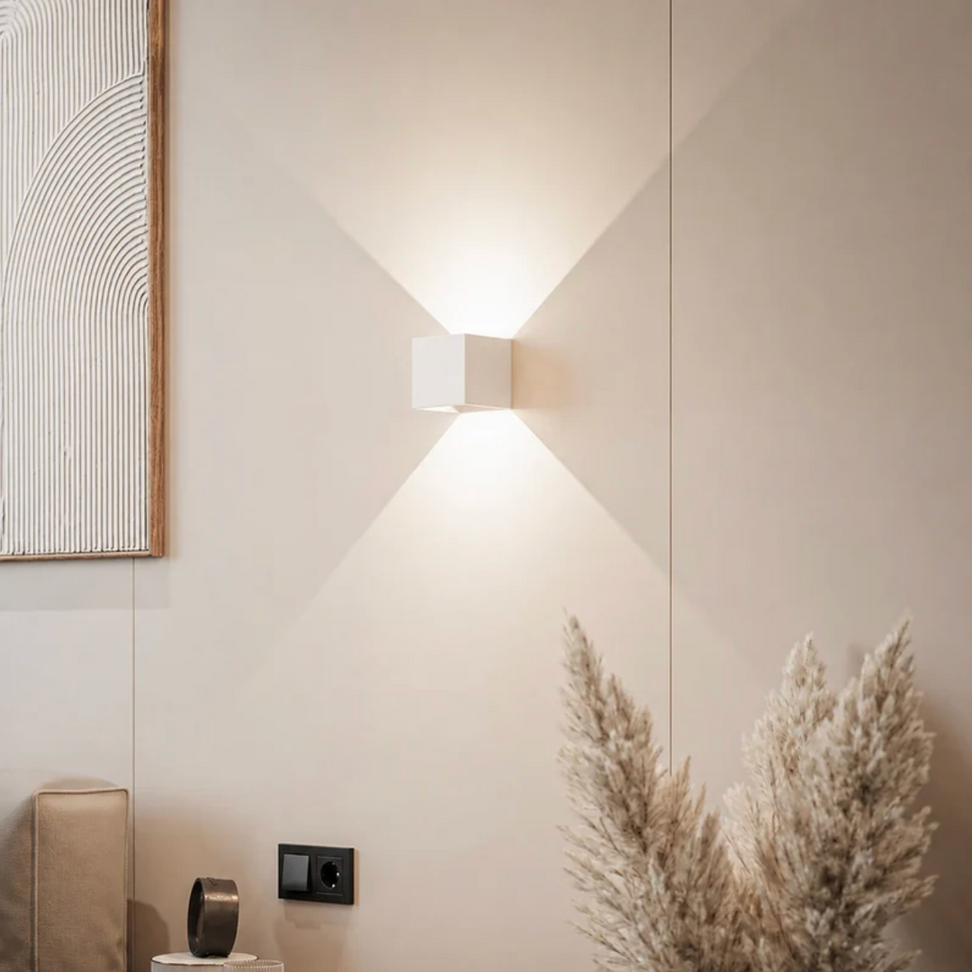 White cube wall lamp casting soft light up and down on a beige wall, with pampas grass and a framed textured artwork. 12