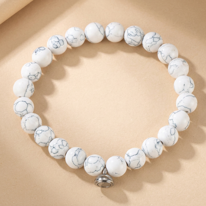 White beaded bracelet with gray marbled veining and a silver magnetic charm, shown flat on a tan background 3
