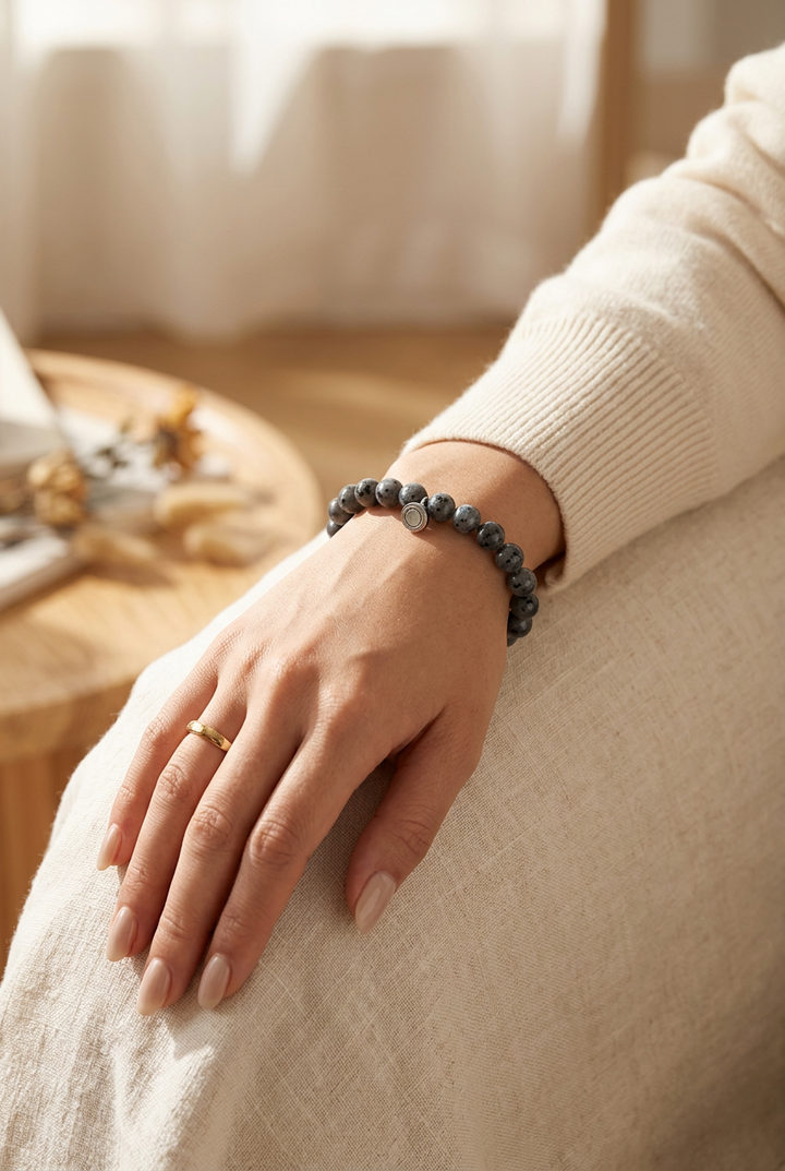 Woman's wrist wearing the gray speckled beaded bracelet with a small silver charm, hand resting on a linen-covered lap 1