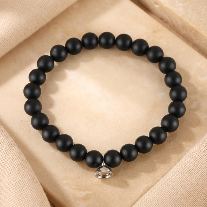 Matte black beaded stretch bracelet with a small silver magnetic charm, laid flat on a beige stone tile 2