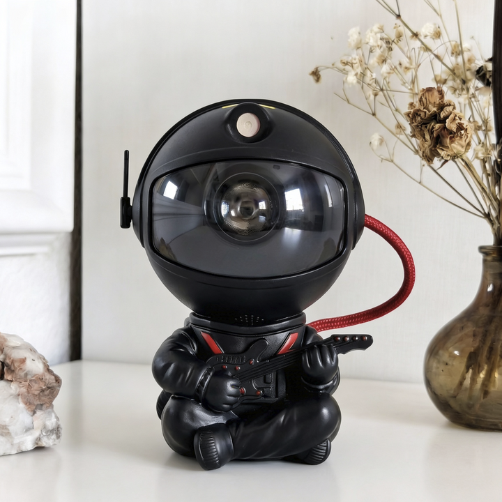 Black astronaut projector sitting cross-legged and playing a guitar, with red accents and a red braided cord, front view.