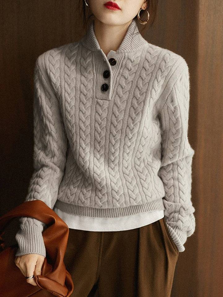 Model in the light gray cable-knit button sweater layered over a white top, holding a tan leather bag against a wood-paneled wall. 14