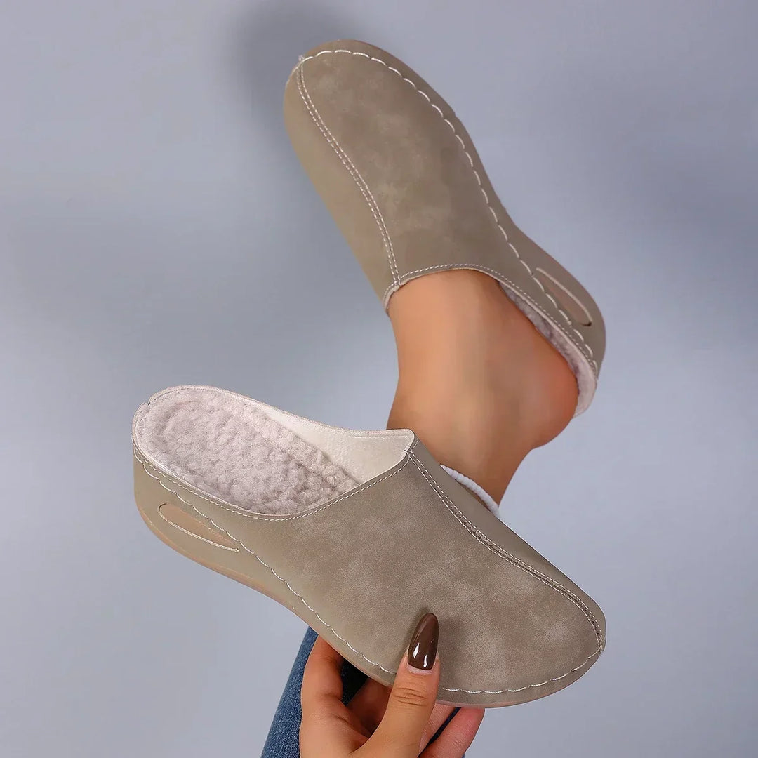 Taupe clog slippers held up in a hand with brown nails and the fluffy fleece lining and stitched edges.