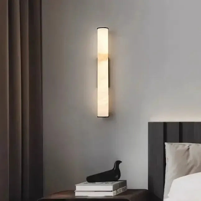 Slim marble sconce glowing above a dark nightstand with a black bird figurine and books, beside a dark headboard and drapes.