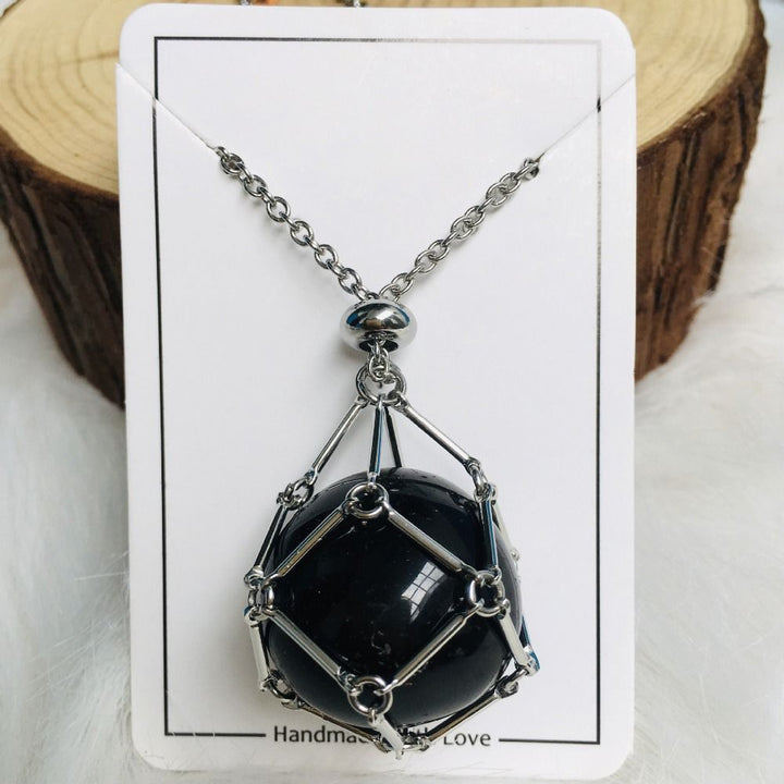 Silver cage necklace with a round glossy black stone on a white card with a wood log slice behind. 8