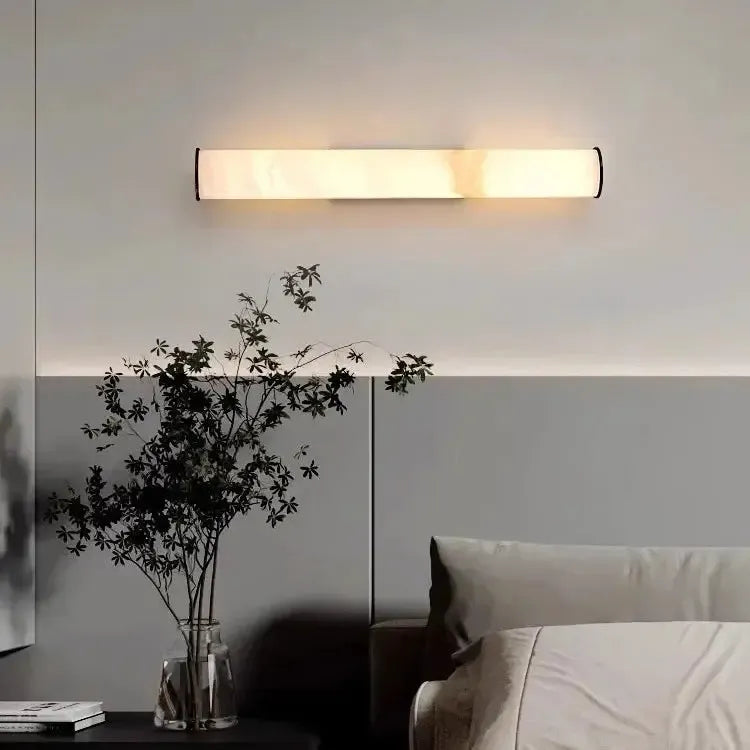 Marble light mounted horizontally on a two-tone gray wall above a bed, with a glass vase of dark branches on the side table.