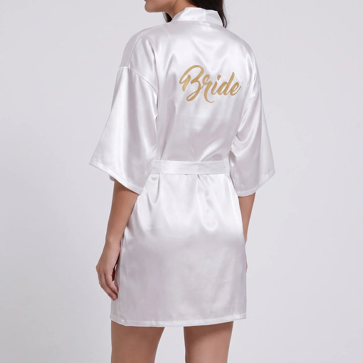 Back view of a model wearing the white satin robe with gold glitter script lettering across the upper back, belt tied at the waist. 3