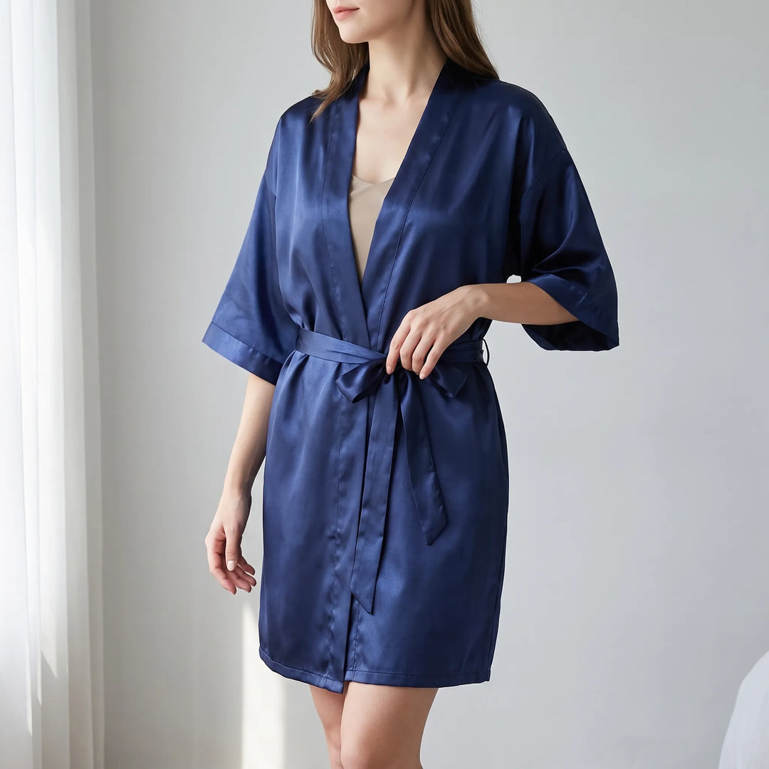 Woman in a navy satin kimono-style wrap robe, belted at the waist, standing beside a sheer curtain in a bright room. 1