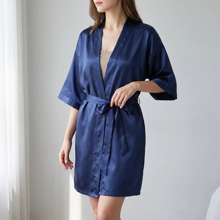 Woman in a navy satin kimono-style wrap robe, belted at the waist, standing beside a sheer curtain in a bright room. 1