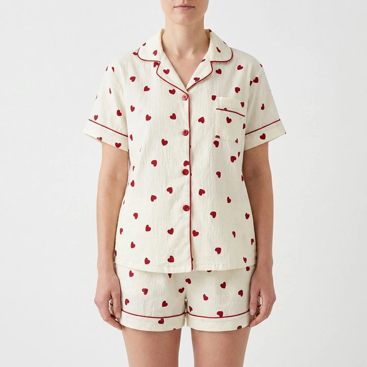 Front view of a model wearing the short-sleeve button-up pajama top and matching shorts in cream with red hearts and red piping. 2