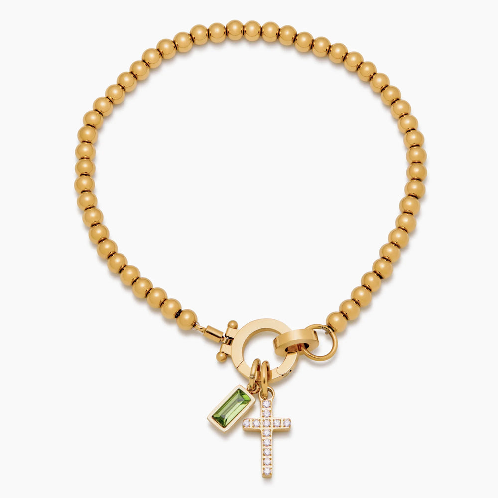 Gold-tone bead bracelet detail with a round clasp, green baguette stone charm and crystal cross charm.