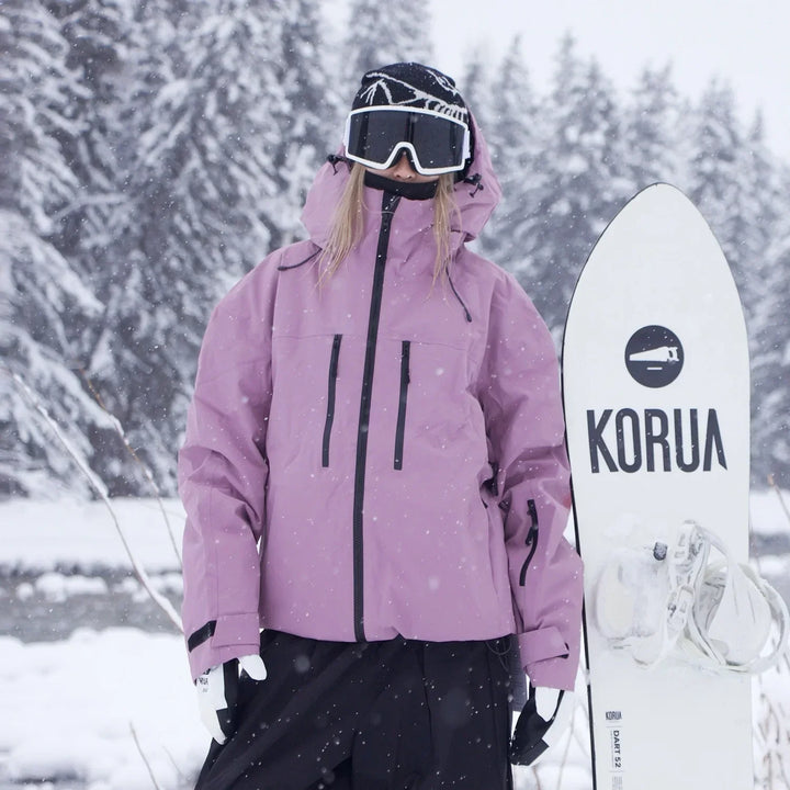 Model in a dusty pink hooded jacket with black chest zippers stands beside a white snowboard in falling snow.