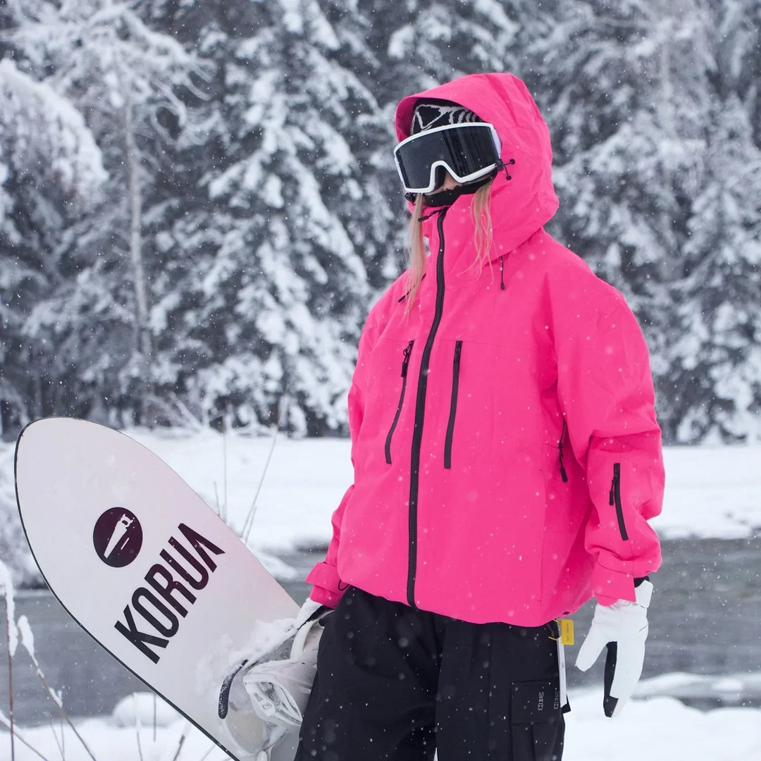 Model in a bright pink hooded jacket with black zippers carries a snowboard past snow-covered evergreens.