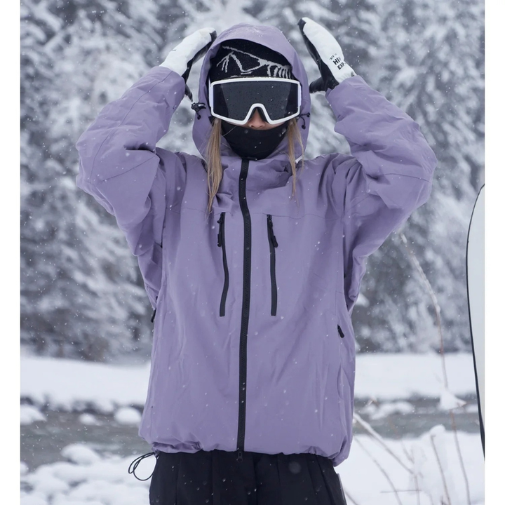 Model in a lavender hooded jacket with black zippers raises both hands to the hood amid falling snow and snowy trees.