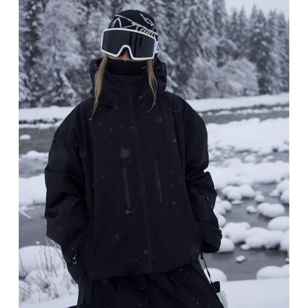 Model wearing the black version of the oversized hooded jacket, hands in pockets, standing by a snowy stream.