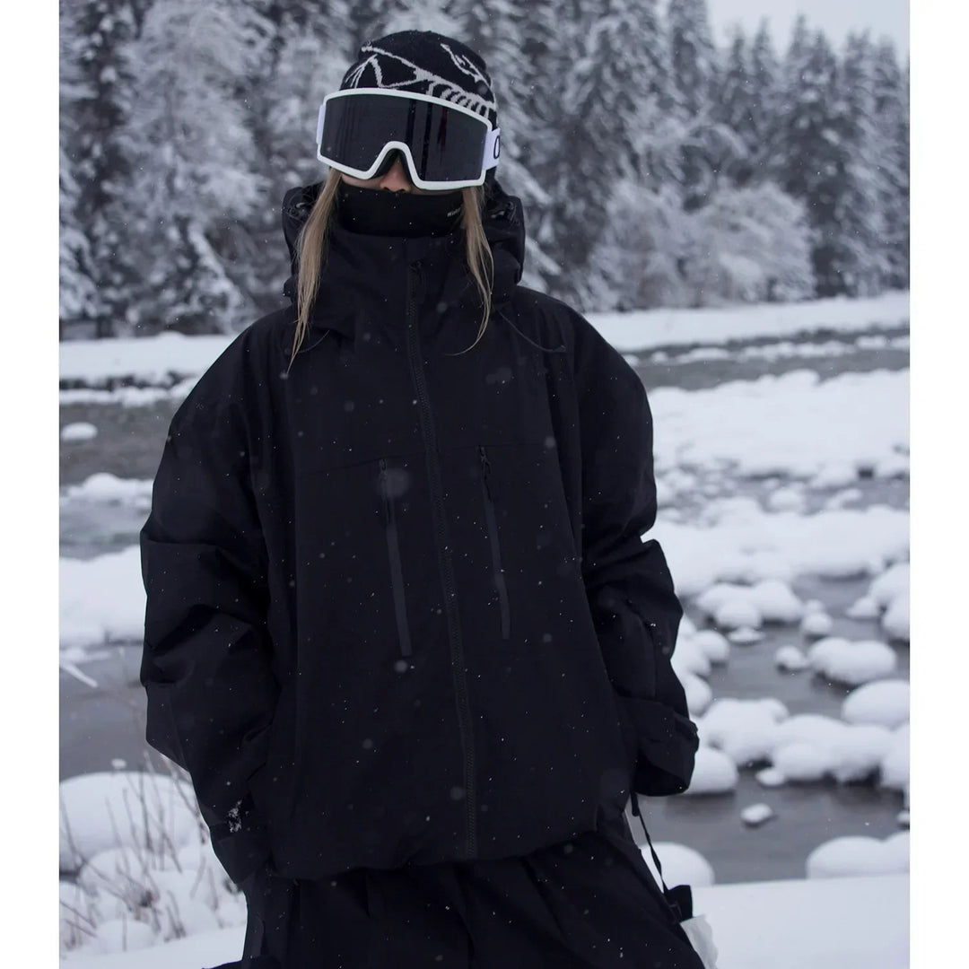 Model wearing the black version of the oversized hooded jacket, hands in pockets, standing by a snowy stream.