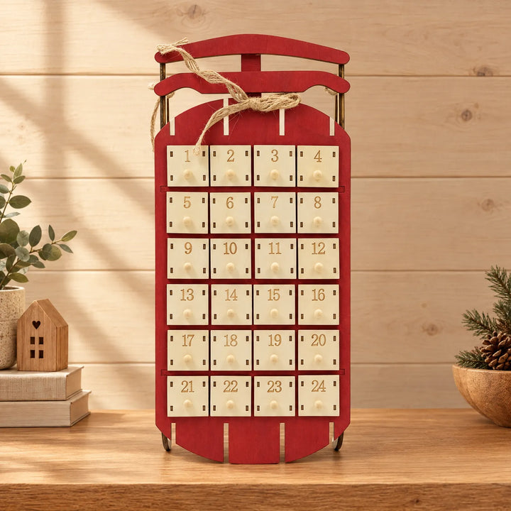 Red wooden sled advent calendar with 24 numbered drawers and a jute bow, standing on a shelf beside books, a plant and pine. 1