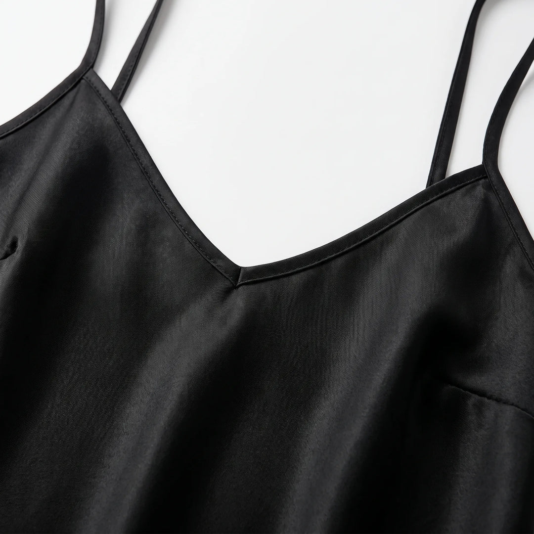 Close-up detail of the black satin slip dress's V-neckline, stitched edging and thin spaghetti straps on a white surface. 5