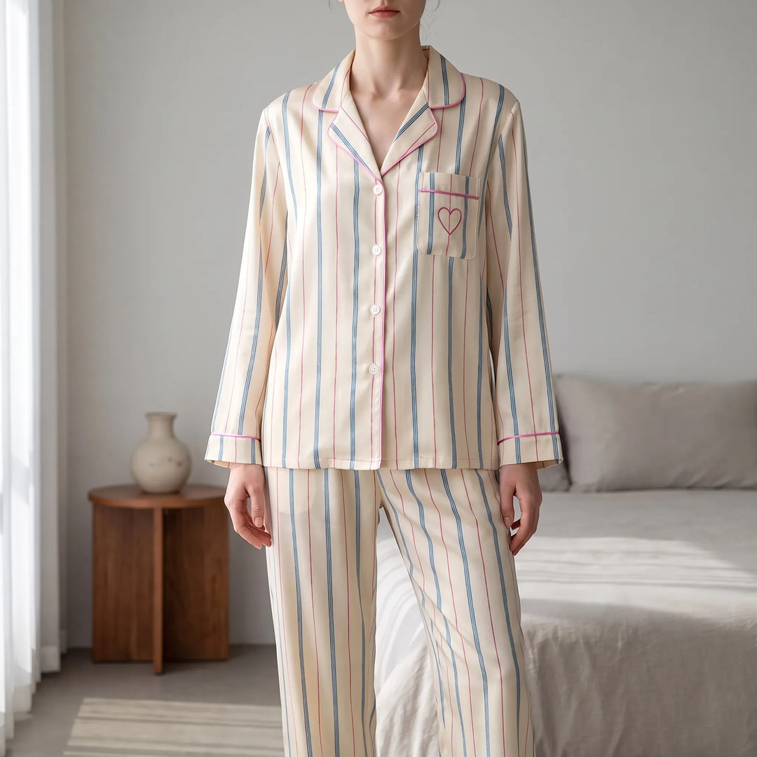 Woman wearing the cream pajama set with blue and red stripes and pink piping, standing in a bedroom, cropped at the face. 1