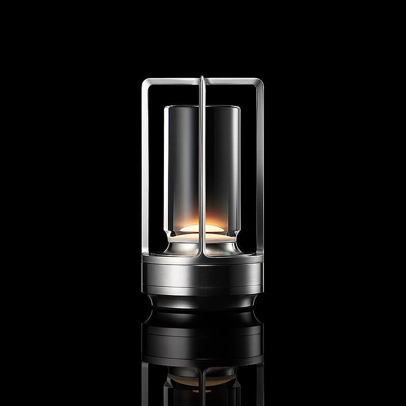 Silver-finish lamp with open frame and clear glass cylinder, softly lit against a black background with reflection.