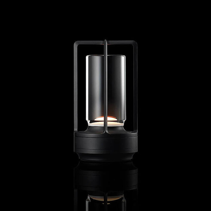 Black-finish lamp with open frame and clear glass cylinder, softly lit against a black background with reflection.