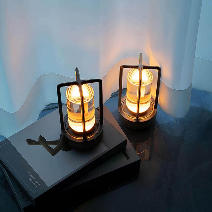 Two dark-framed lamps with warm glowing cylinders sit on stacked books beside a sheer curtain, lit in a bluish room.