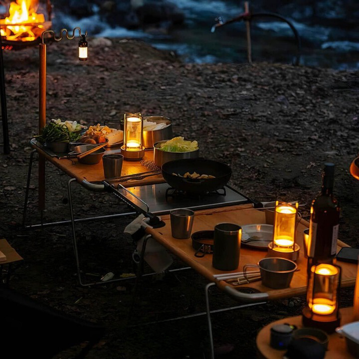 Several lamps glowing amber on camping tables set with cookware and food at dusk near a campfire.