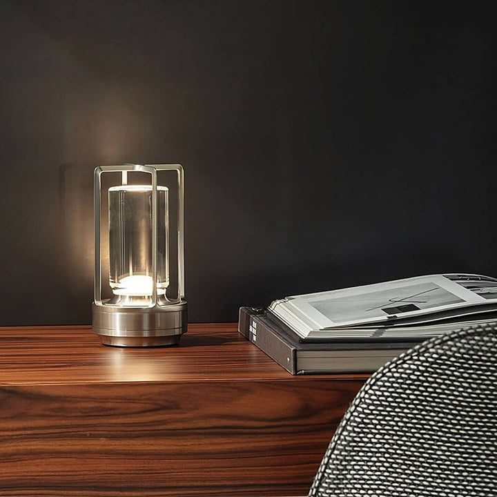 Silver-finish lamp lit on a wooden sideboard beside an open book, against a dark wall.